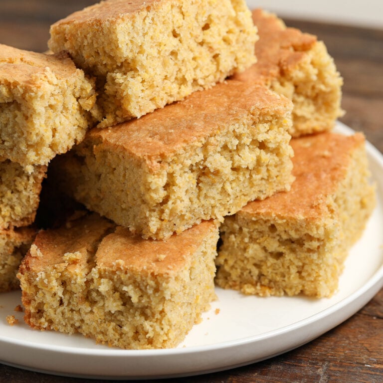 A plate stacked with several pieces of golden-brown gluten free cornbread squares, showing a moist, crumbly texture on a wooden table.