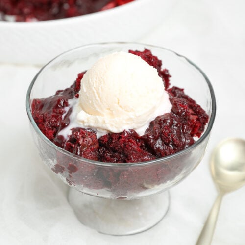 A glass bowl filled with a warm red velvet pudding cake with a scoop of vanilla ice cream, next to a silver spoon on a white surface.