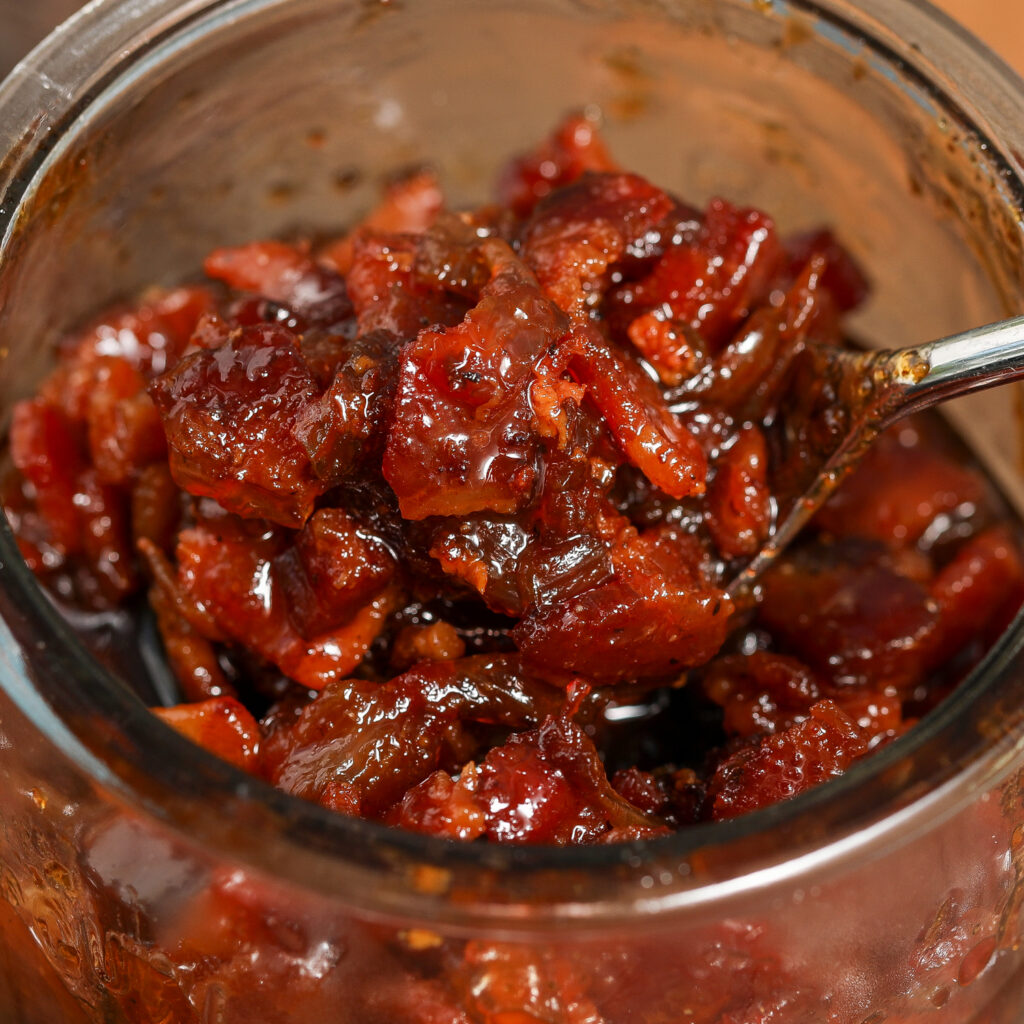 Close-up of a jar filled with chunky, glossy BBQ Bacon Jam, with a spoon scooping out the rich, caramelized mixture.