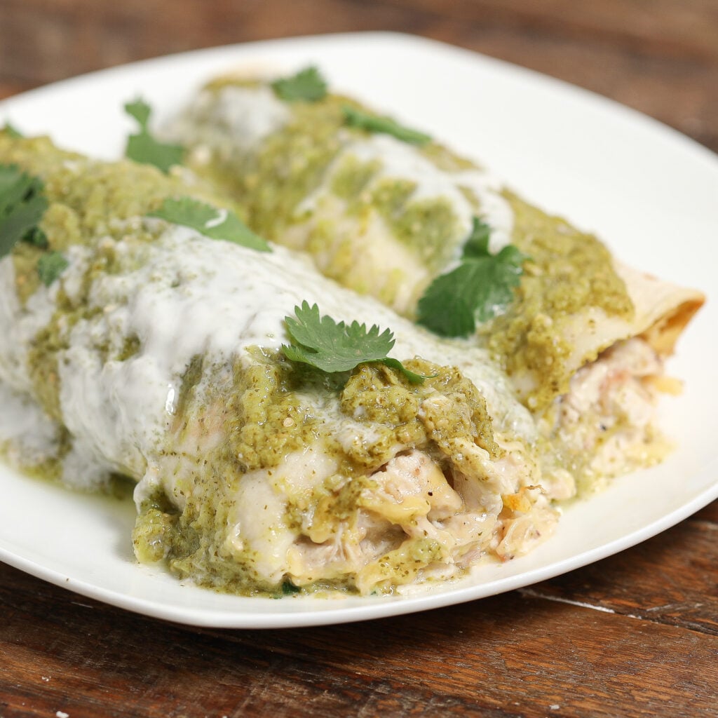 Verde Chicken Enchiladas: Two chicken enchiladas topped with green salsa, creamy white sauce, and fresh cilantro, served on a white plate on a wooden table.