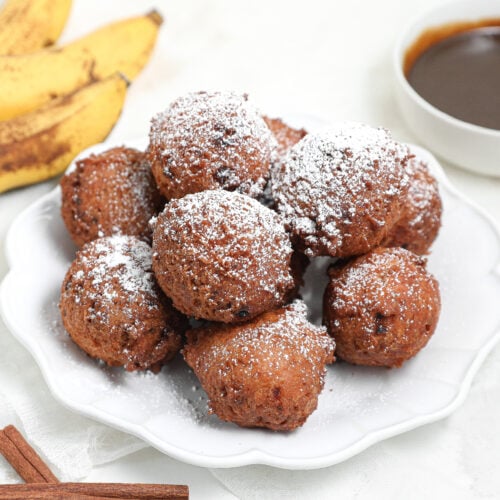 A plate of powdered sugar-dusted banana fritters is shown, with ripe bananas, a bowl of chocolate sauce, and cinnamon sticks in the background, all on a white surface.