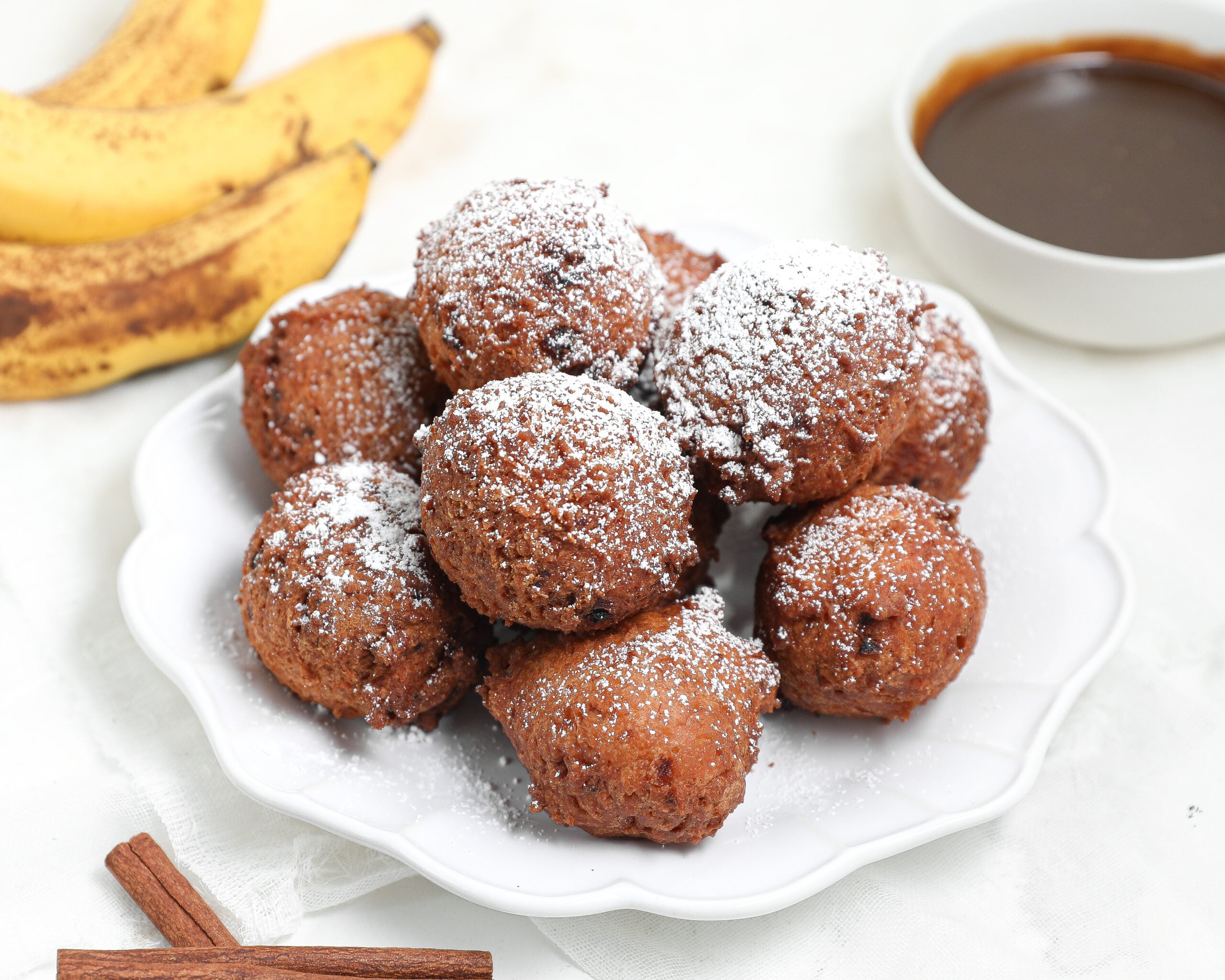 A plate of powdered sugar-dusted banana fritters is shown, with ripe bananas, a bowl of chocolate sauce, and cinnamon sticks in the background, all on a white surface.