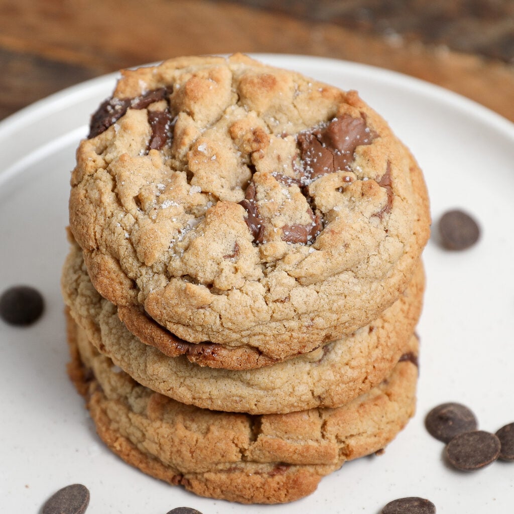 A stack of three browned butter cookies with chocolate chips sits on a white plate, with a few chocolate chips scattered around. The cookies look golden brown and soft, with visible chocolate chunks.