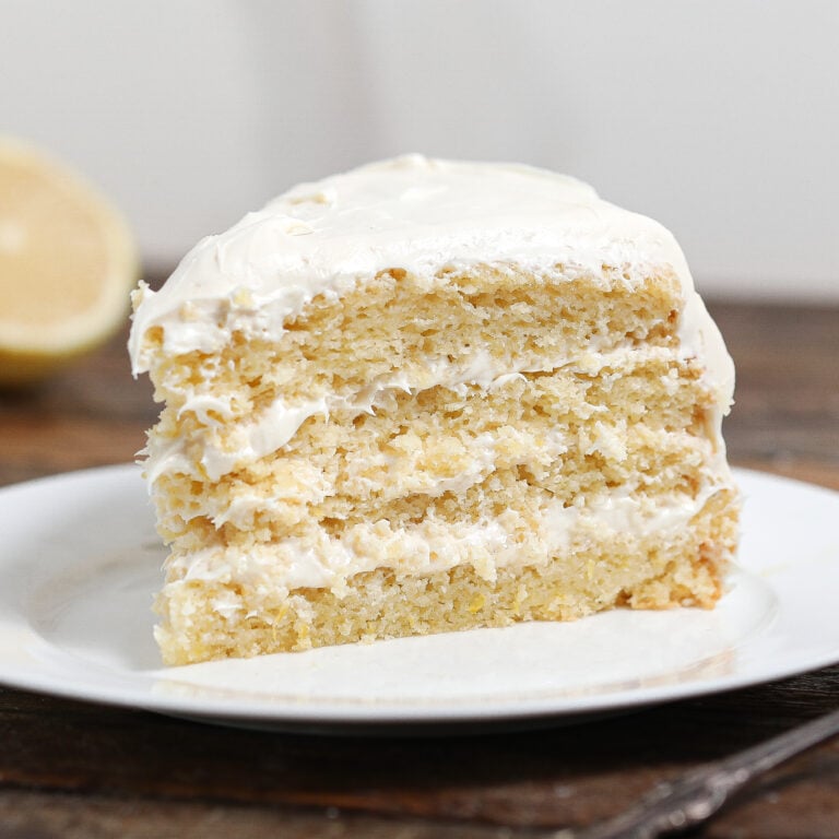 A slice of three-layer Lemon Cake with Cream Cheese Frosting between each layer and on top, served on a white plate. A lemon half is blurred in the background.