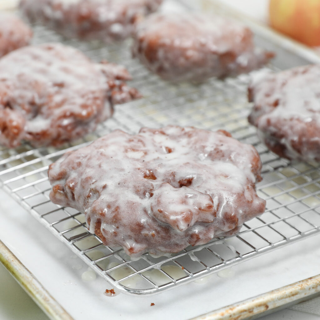 Glazed apple fritters cooling on a wire rack placed over a baking sheet, with icing dripping off the pastries onto the rack and tray below.