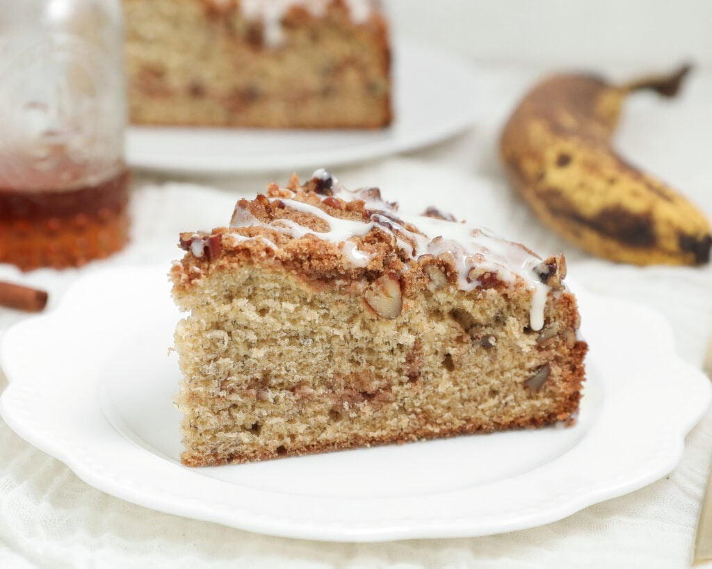 Banana bread coffee cake with cinnamon pecan crumb topping and vanilla glaze served on a white plate.