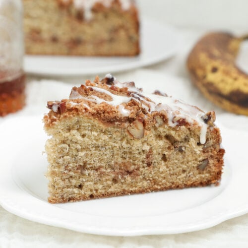 Banana bread coffee cake with cinnamon pecan crumb topping and vanilla glaze served on a white plate.
