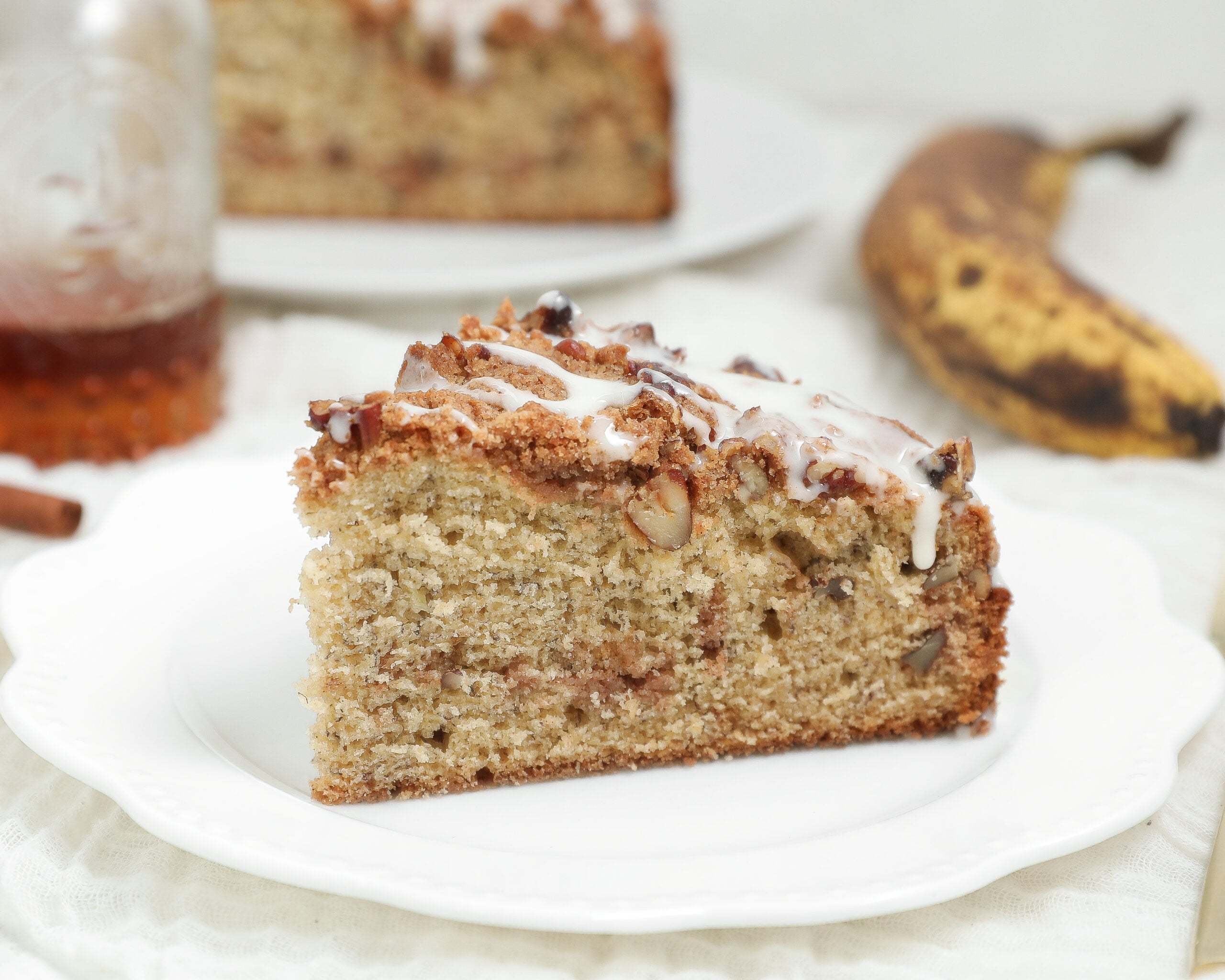 Banana bread coffee cake with cinnamon pecan crumb topping and vanilla glaze served on a white plate.