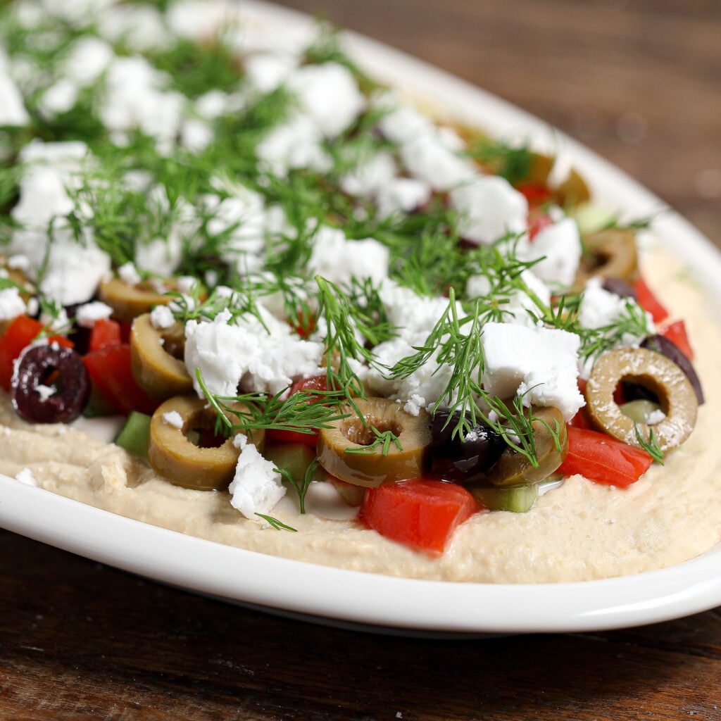 A close-up of a plate with 7-Layer Greek Dip featuring hummus topped with chopped tomatoes, sliced green and black olives, crumbled feta cheese, and fresh dill, served on a white dish.
