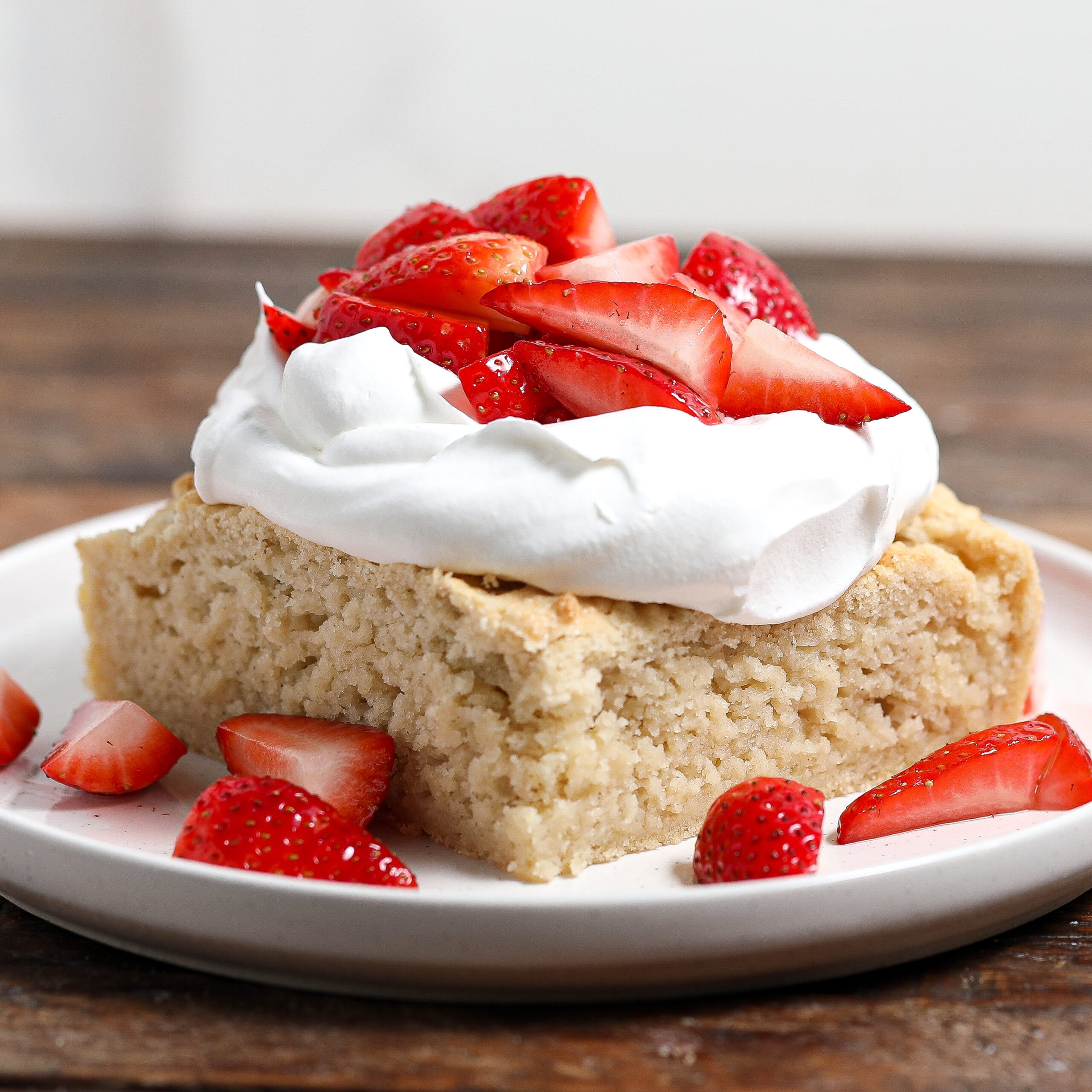 A slice of biscuit cake shortcake topped with a generous dollop of whipped cream and fresh sliced strawberries, served on a white plate with extra strawberry pieces around it.