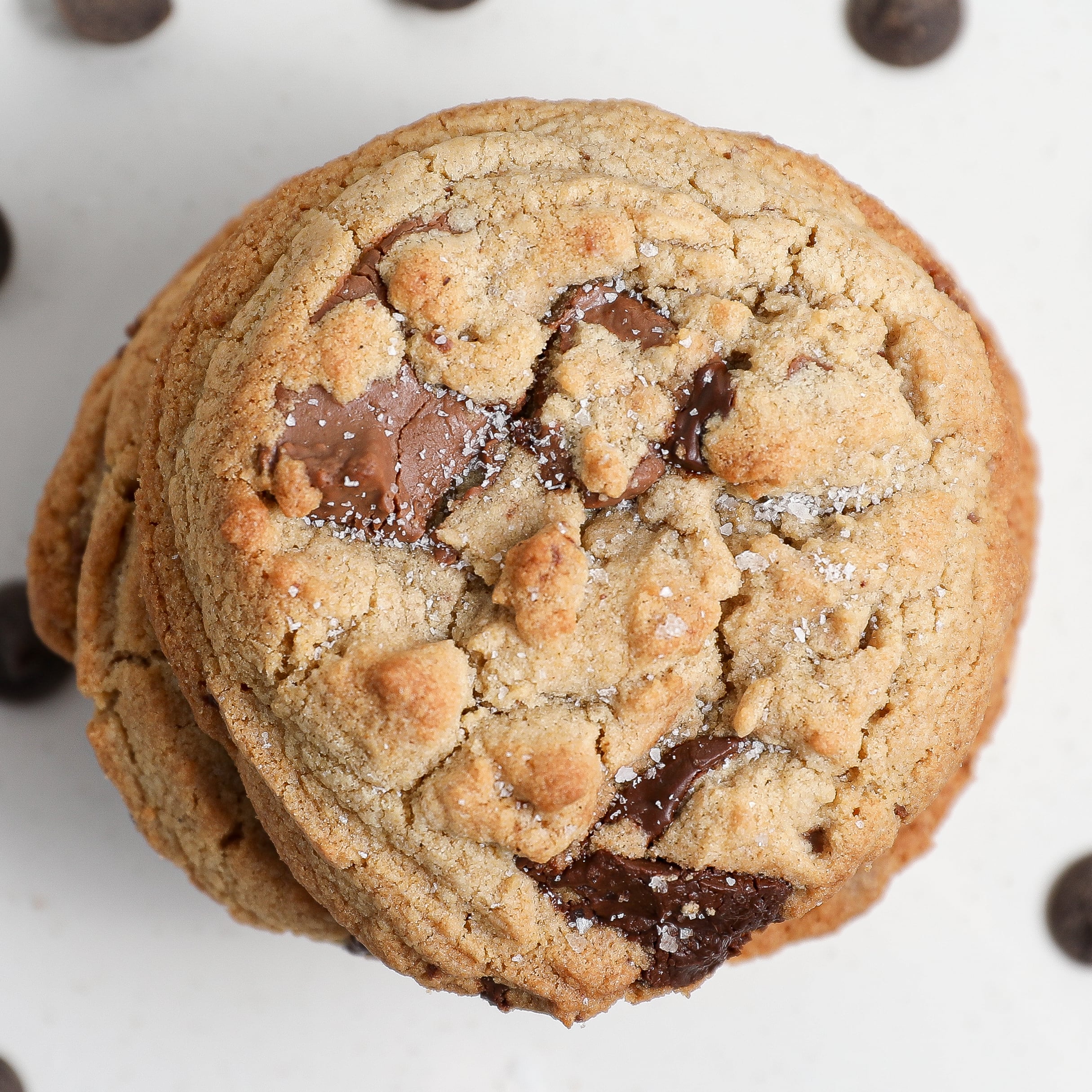 A close-up, top-down view of a stack of browned butter chocolate chip cookies on a white surface, sprinkled with a bit of salt, with a few chocolate chips scattered around.