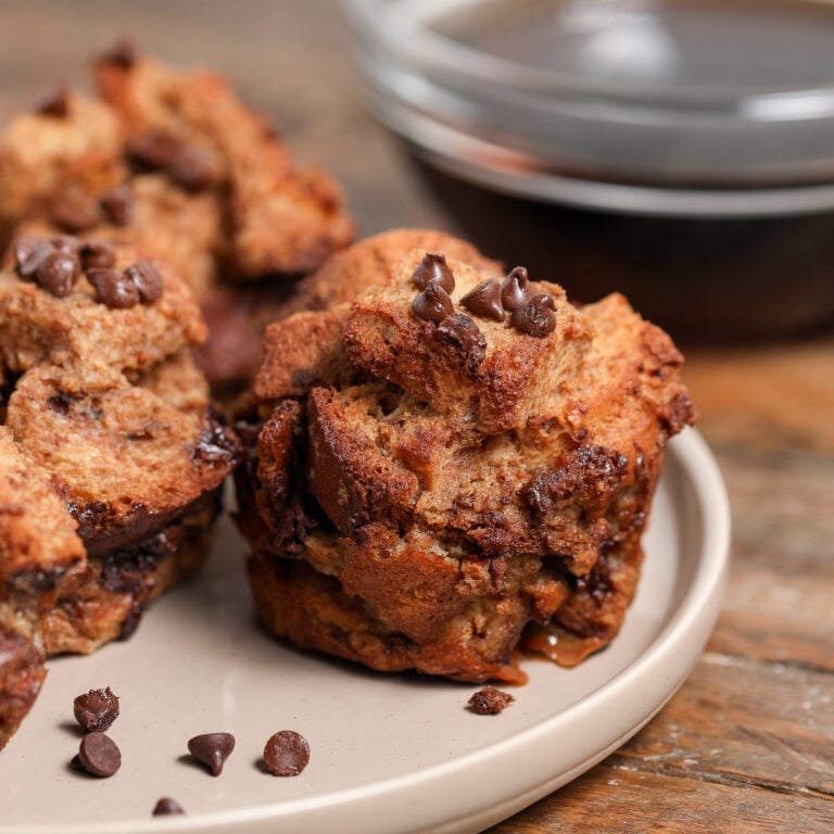 A close-up of Chocolate Chip French Toast Bites and bread pudding muffins on a beige plate, with extra chocolate chips scattered around and a bowl of syrup in the background on a wooden table.