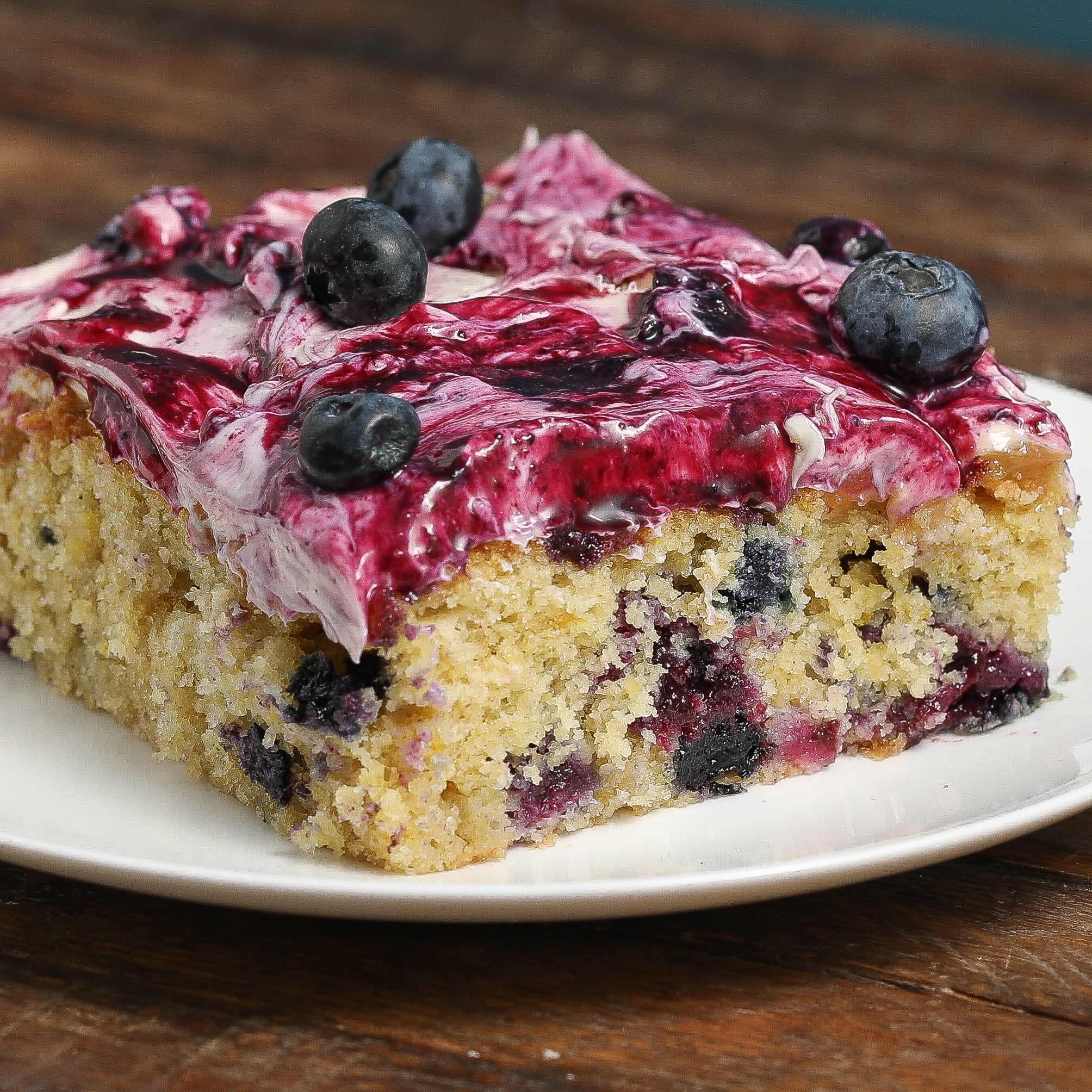 A slice of Lemon Blueberry Poke Sheet Cake topped with creamy purple frosting and fresh blueberries is served on a white plate, sitting on a rustic wooden surface.