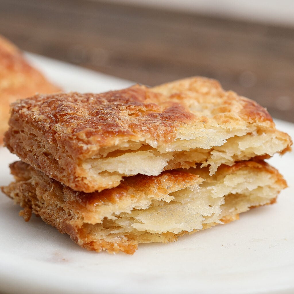 Two pieces of golden, flaky pastry with crispy, layered texture are stacked on a white surface. The pastries have a slightly browned, buttery crust and soft layers inside—perfect for pairing with fresh strawberry sauce.