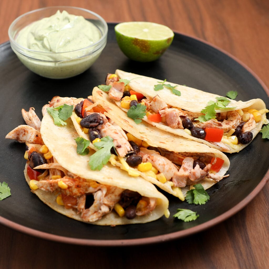 Enjoy our Southwestern Chicken Tacos Recipe: three soft tacos filled with shredded chicken, black beans, corn, diced red peppers, and cilantro. Served on a black plate with a lime wedge and creamy green sauce.
