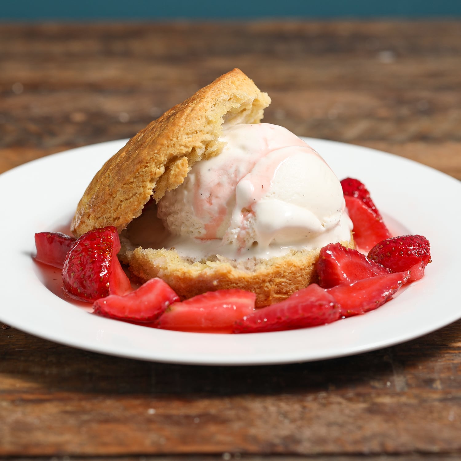 A scoop of vanilla ice cream is sandwiched in a buttermilk biscuit and served on a white plate, surrounded by sliced strawberries in syrup—a delightful twist on Strawberry Shortcake with Buttermilk Biscuits, all on a rustic wooden table.