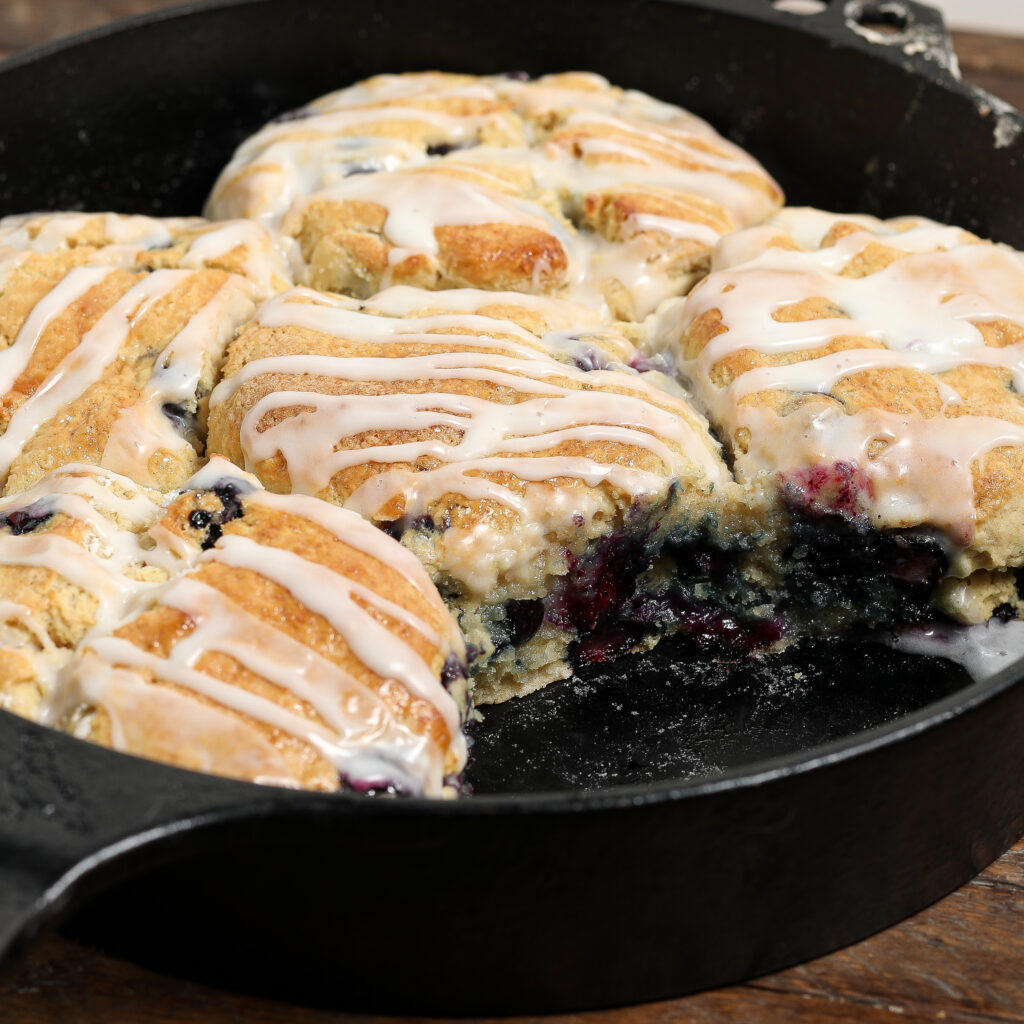 A close-up of glazed blueberry scones, reminiscent of blueberry biscuits, in a cast iron skillet, with one scone partially removed to reveal a soft, moist interior studded with blueberries.
