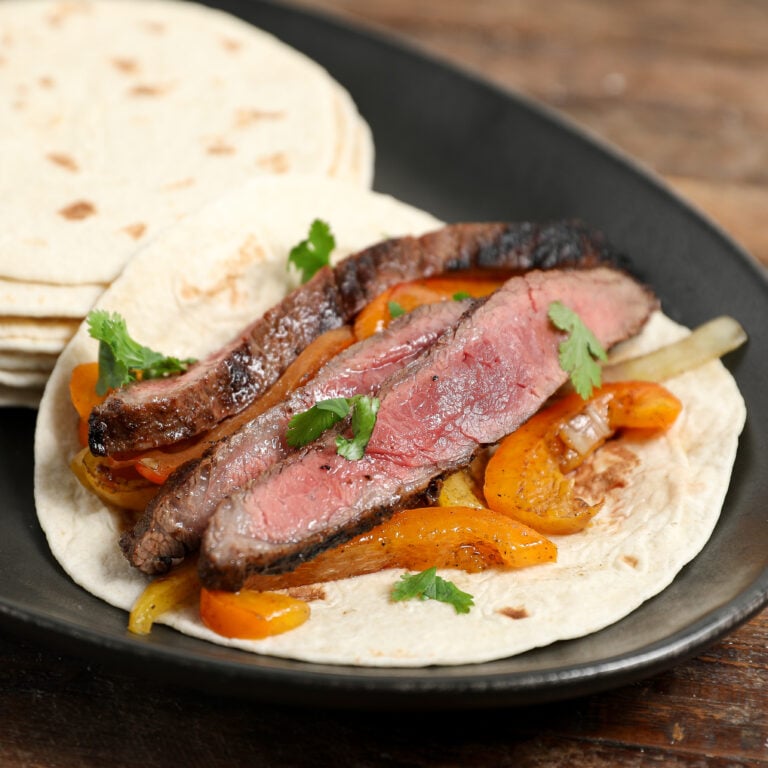 A flour tortilla topped with grilled steak slices, sautéed orange bell peppers, onions, and fresh cilantro, this Steak Fajitas Recipe is served on a black plate with extra tortillas in the background.