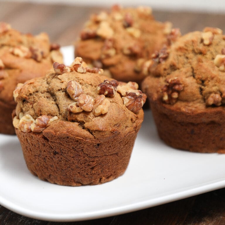 Four homemade banana nut muffins topped with walnuts are arranged on a white plate, with a rustic wooden surface in the background.