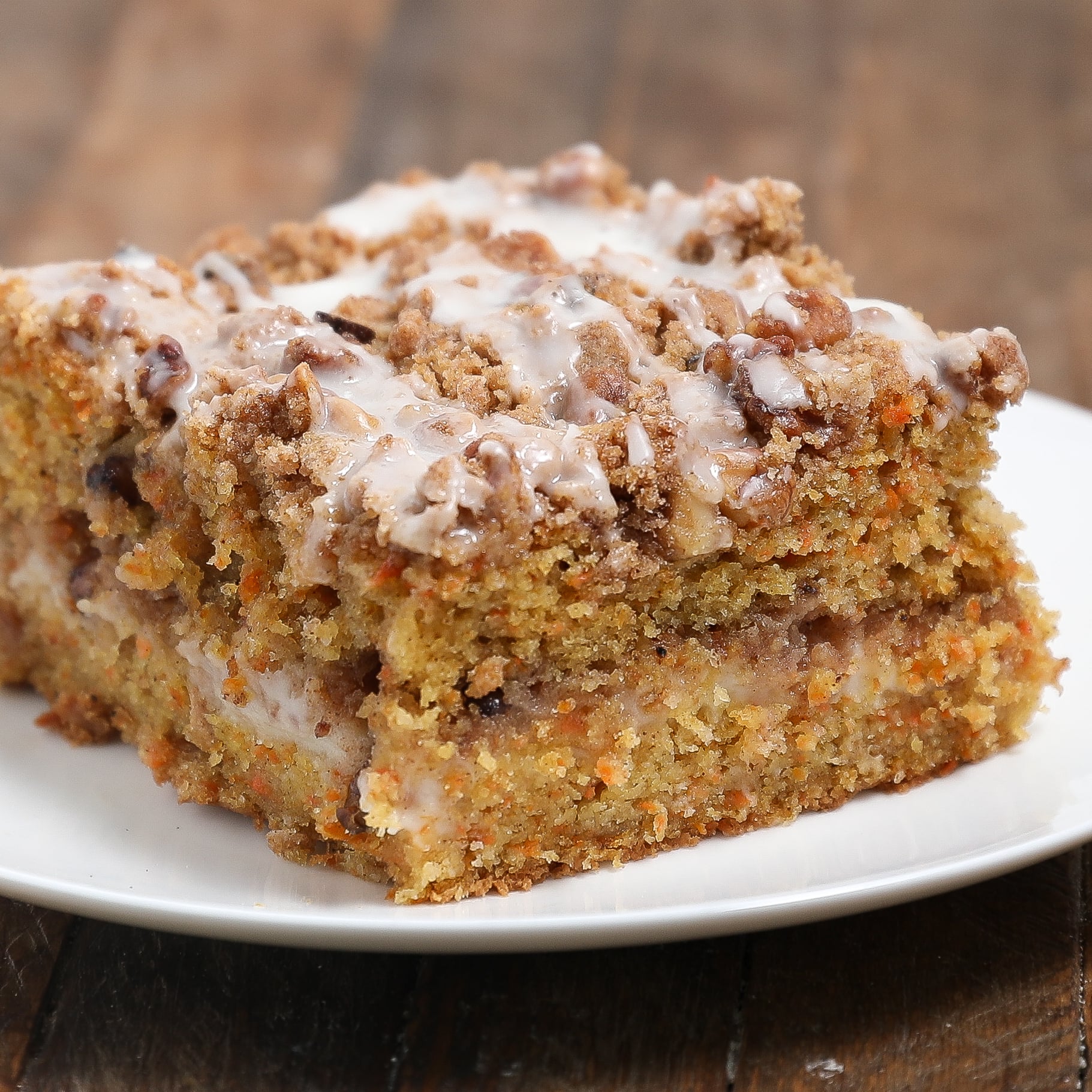 A slice of moist Carrot Cake Coffee Cake topped with crumbly streusel and drizzled with white icing, served on a white plate atop a wooden surface.