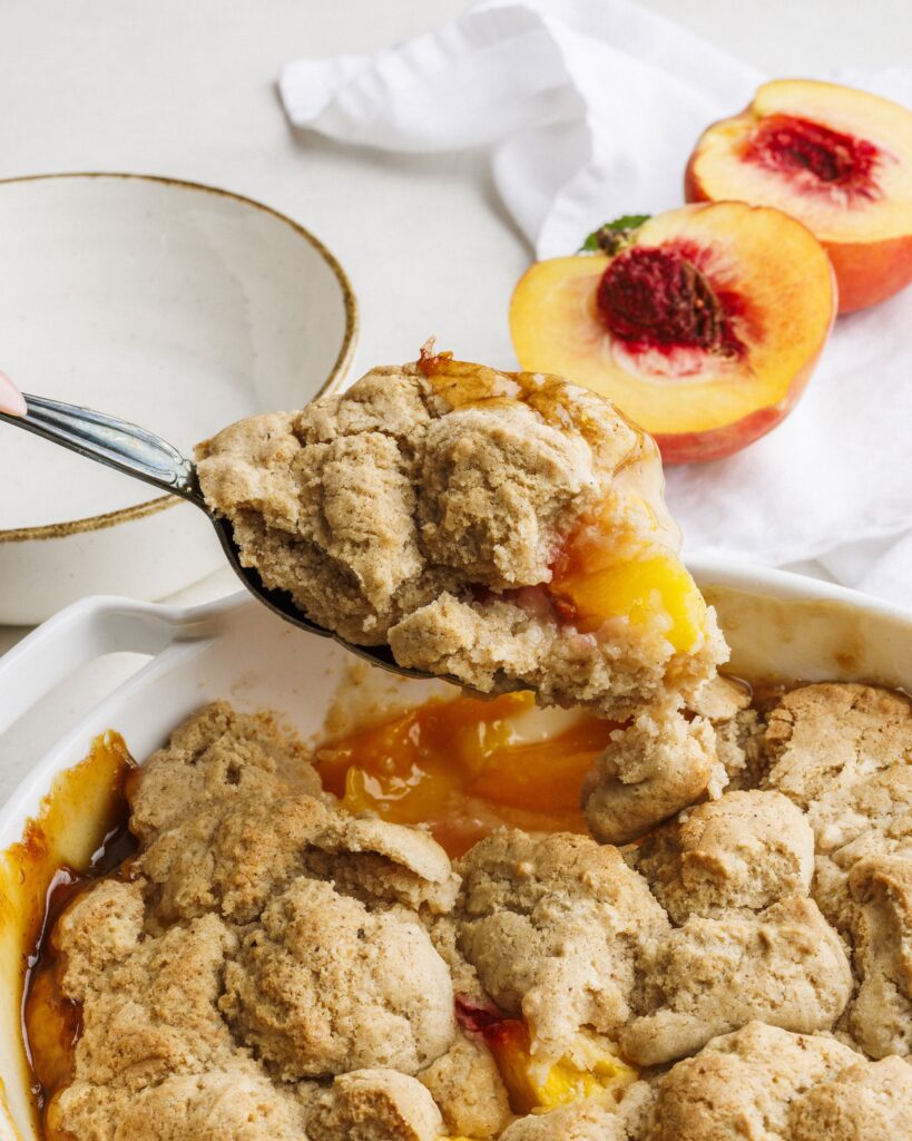 A spoon scoops a portion of peach cobbler from a baking dish, with juicy peaches and golden-brown biscuit topping—just like the classic recipe from the Salty Cooker Cookbook. In the background, a halved peach and an empty bowl are visible.