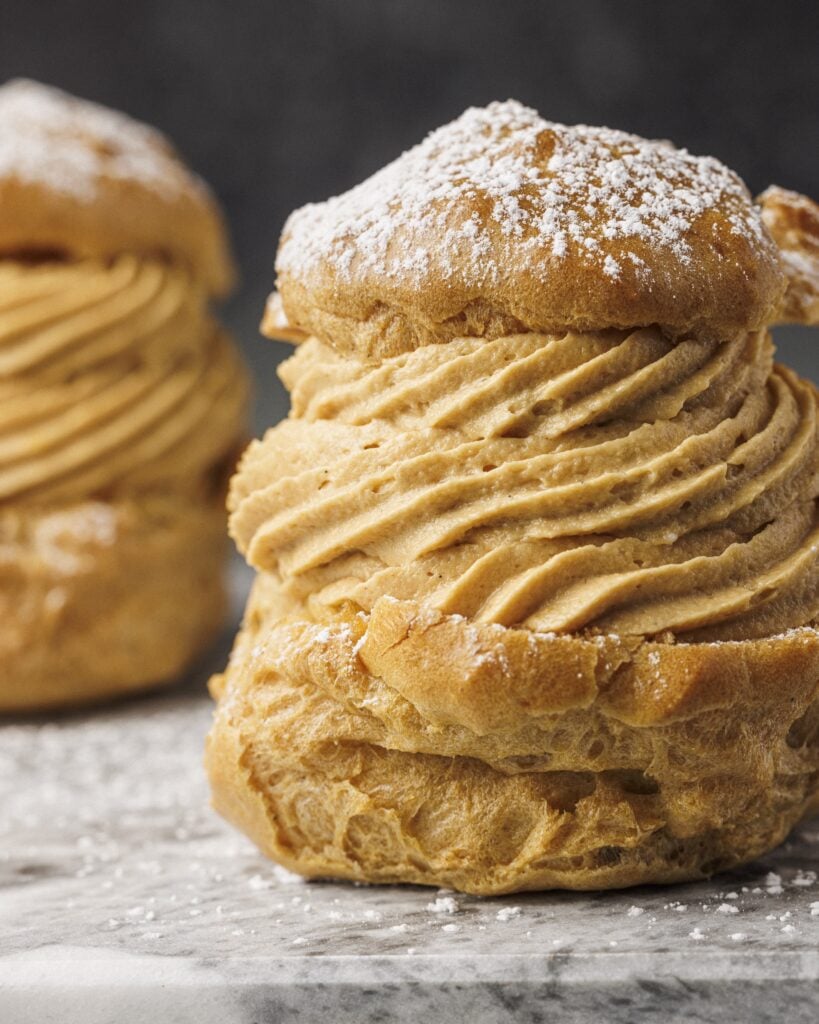 A close-up of a cream puff filled with swirled coffee-flavored cream and topped with powdered sugar, inspired by recipes from the Salty Cooker Cookbook. Another cream puff is blurred in the background on a marble surface dusted with sugar.
