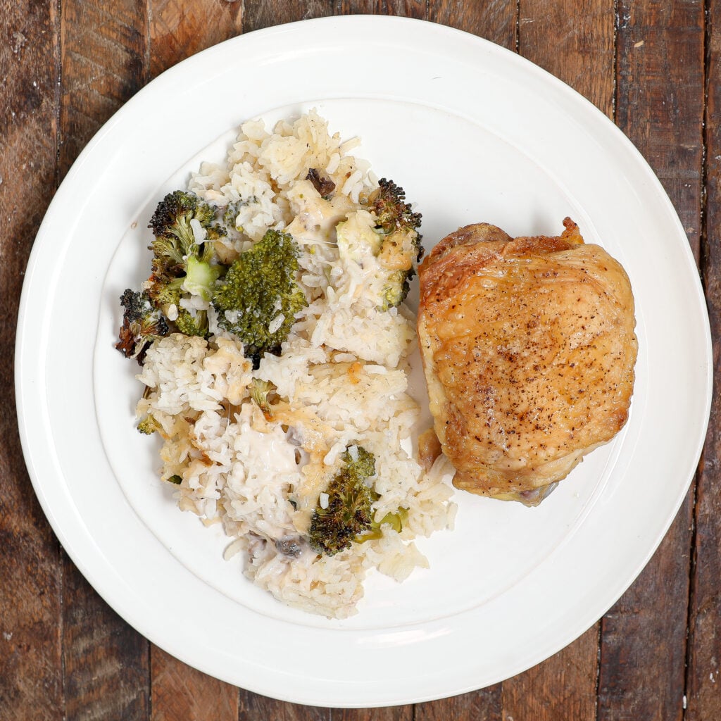 A white plate with a serving of Parmesan Chicken Broccoli Rice Casserole sits next to baked chicken thigh, both placed on a rustic wooden table alongside mixed vegetables.