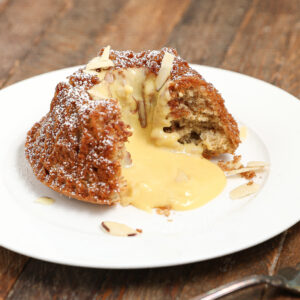A small Almond Cherry Cake with Creme Anglaise sits on a white plate, cut open to reveal creamy yellow custard filling oozing out, topped with powdered sugar and almond slices on a rustic wooden table.