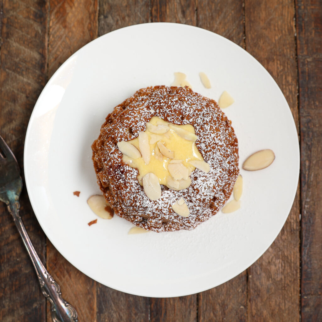 A round Almond Cherry Cake with Creme Anglaise, topped with powdered sugar, sliced almonds, and a drizzle of cream, sits on a white plate on a wooden table, with a spoon beside the plate.