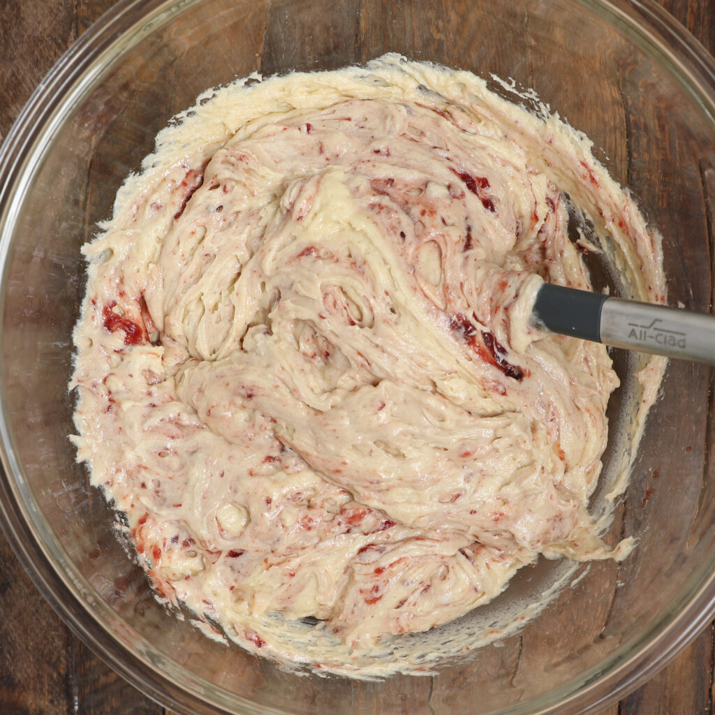 A glass bowl filled with creamy Almond Cherry Cake with Creme Anglaise batter swirled with red fruit or jam, being mixed with a gray spatula, on a wooden surface.