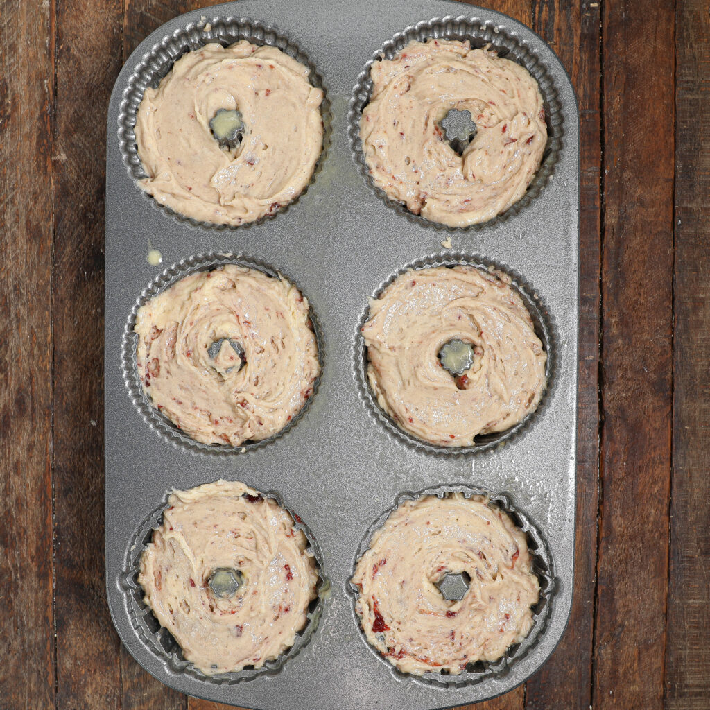 A six-cup donut pan filled with unbaked donut batter, featuring swirls reminiscent of an Almond Cherry Cake with Creme Anglaise, sits on a rustic wooden surface.