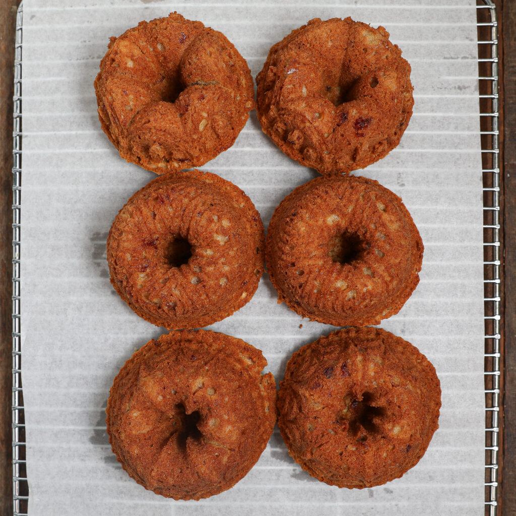 Six small Almond Cherry Cakes with Creme Anglaise arranged in two columns and three rows cool on a wire rack lined with parchment paper. The cakes are golden brown with a slightly rough texture.