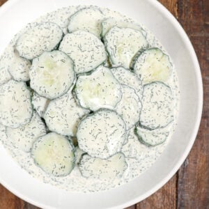 A white bowl filled with cucumber salad—sliced cucumbers coated in a creamy dill sauce—rests on a wooden surface.