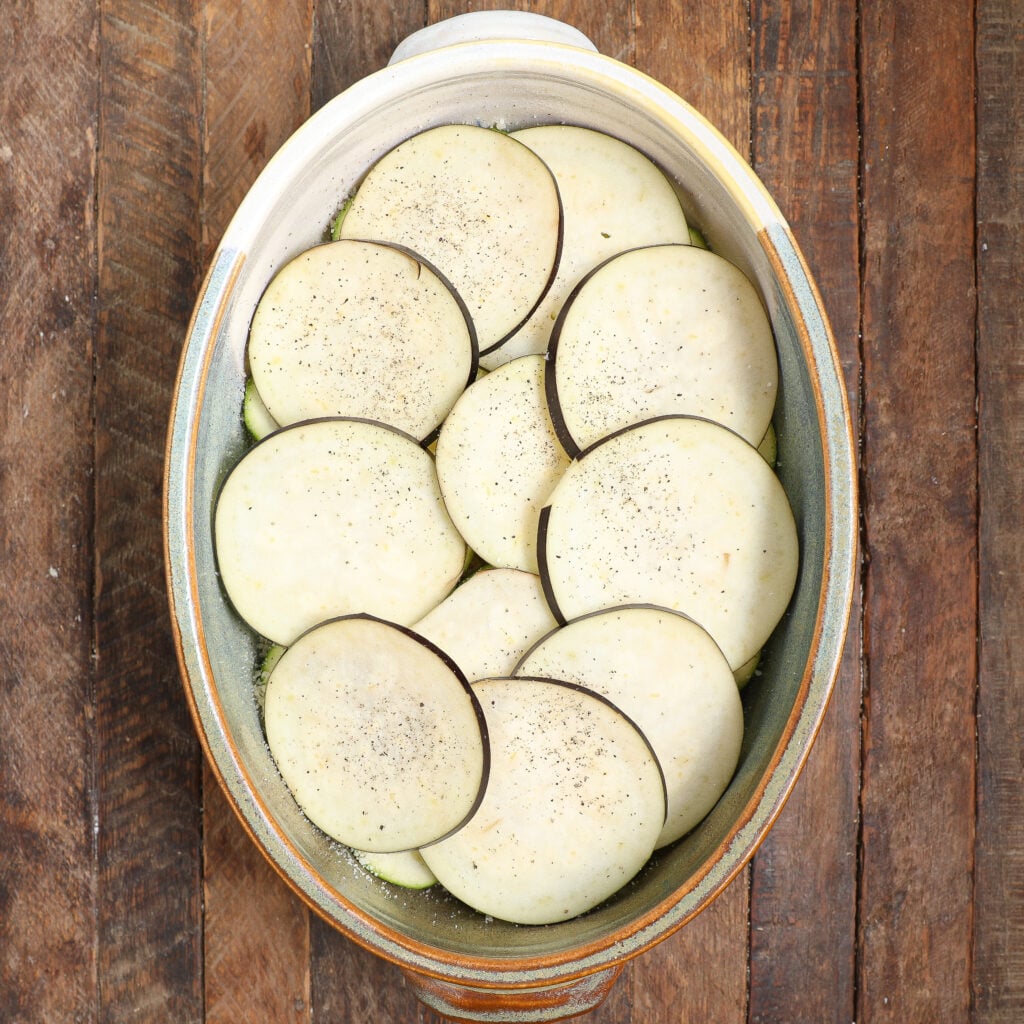 Sliced raw eggplant arranged in an oval ceramic baking dish, sprinkled with black pepper, on a rustic wooden surface—perfect for preparing a Healthier Moussaka.