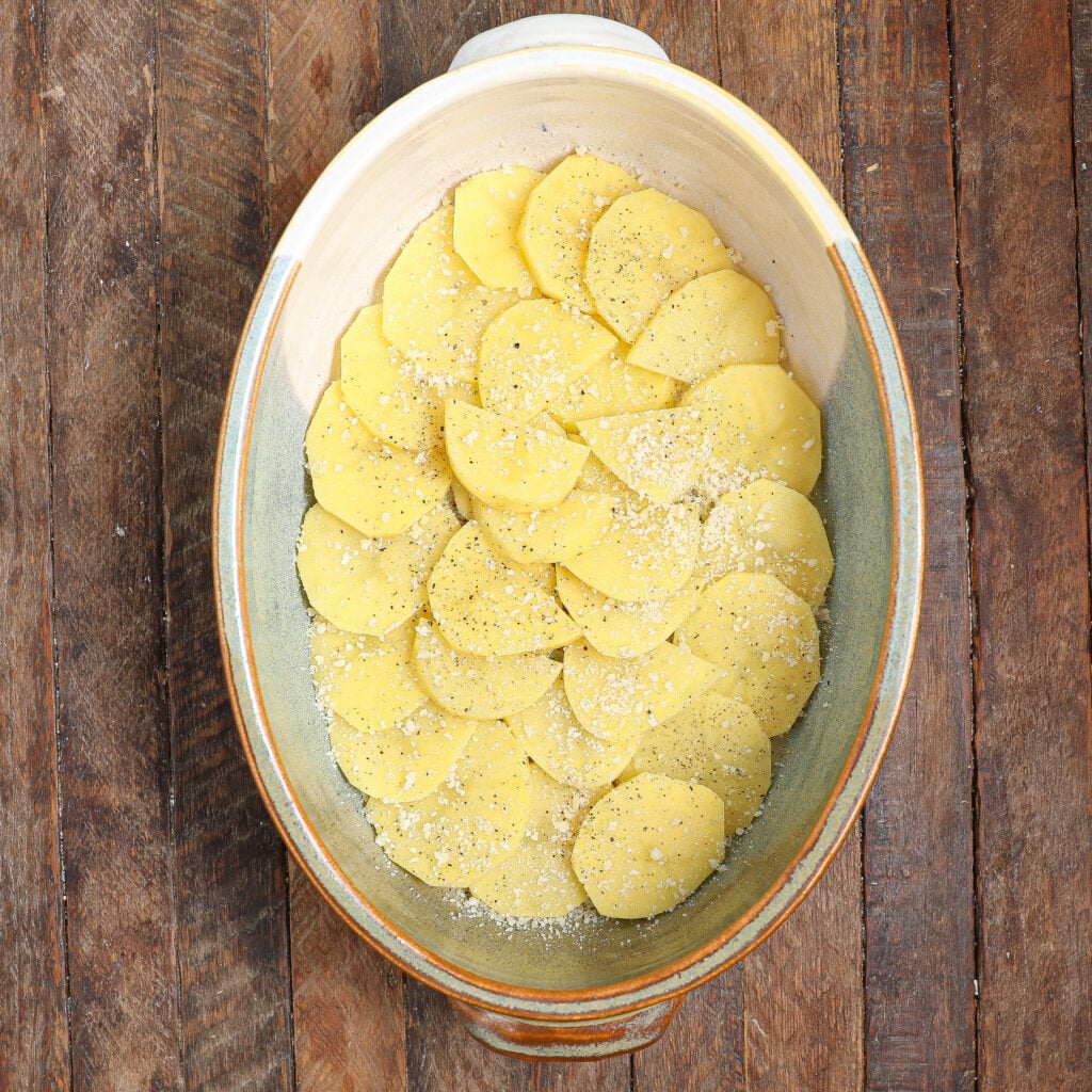 Sliced raw potatoes layered in an oval ceramic baking dish, inspired by a Healthier Moussaka, sprinkled with grated cheese and seasoning, on a rustic wooden surface.