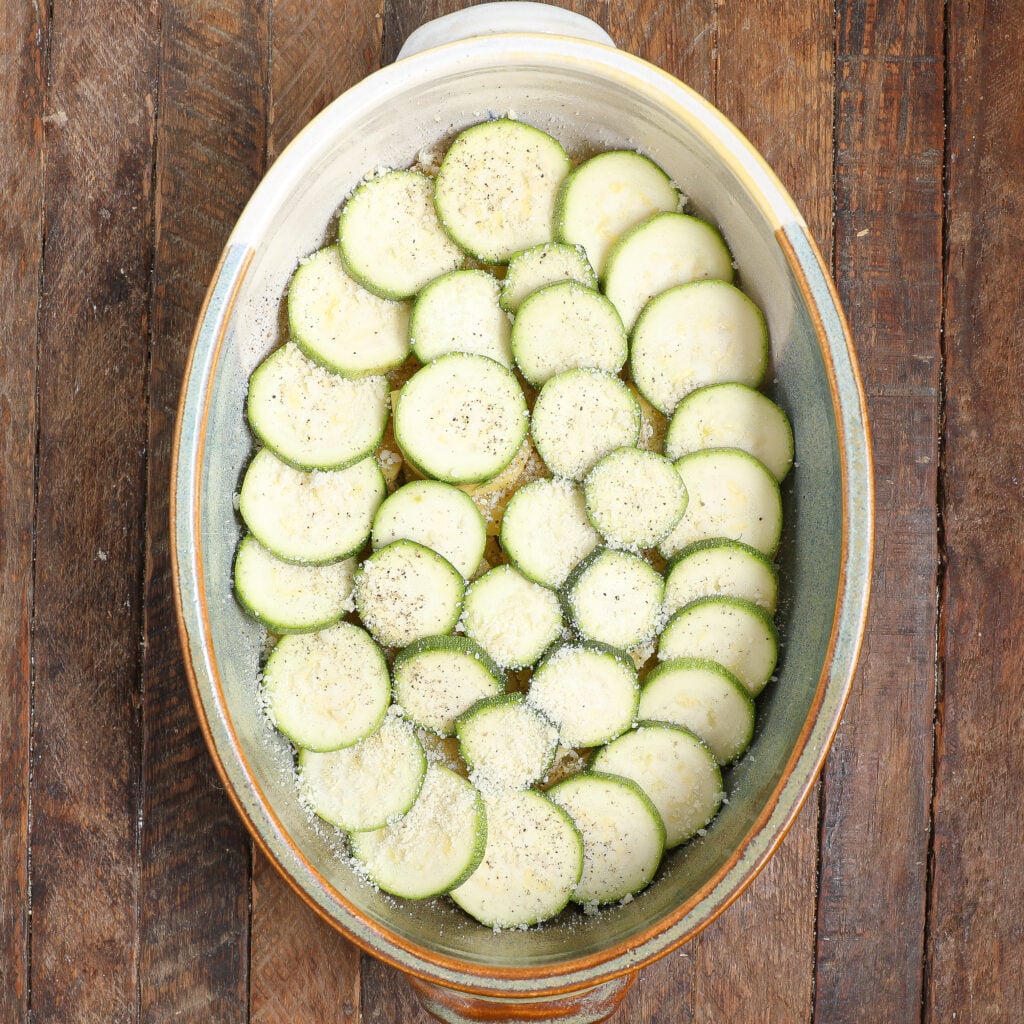 A ceramic oval baking dish filled with sliced zucchini arranged in overlapping layers, sprinkled with grated cheese and black pepper, sits on a rustic wooden surface—a perfect base for a Healthier Moussaka.