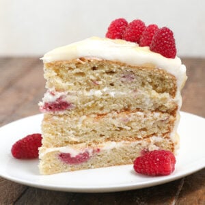 A three-layer slice of Raspberry Lemon Cake with raspberries and cream filling, topped with frosting and fresh raspberries, sits on a white plate on a wooden surface.