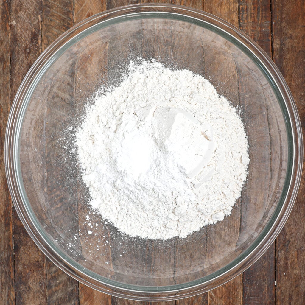 A clear glass bowl filled with white flour and a small mound of baking powder, perfect for making Raspberry Lemon Cake, sits on a wooden surface.