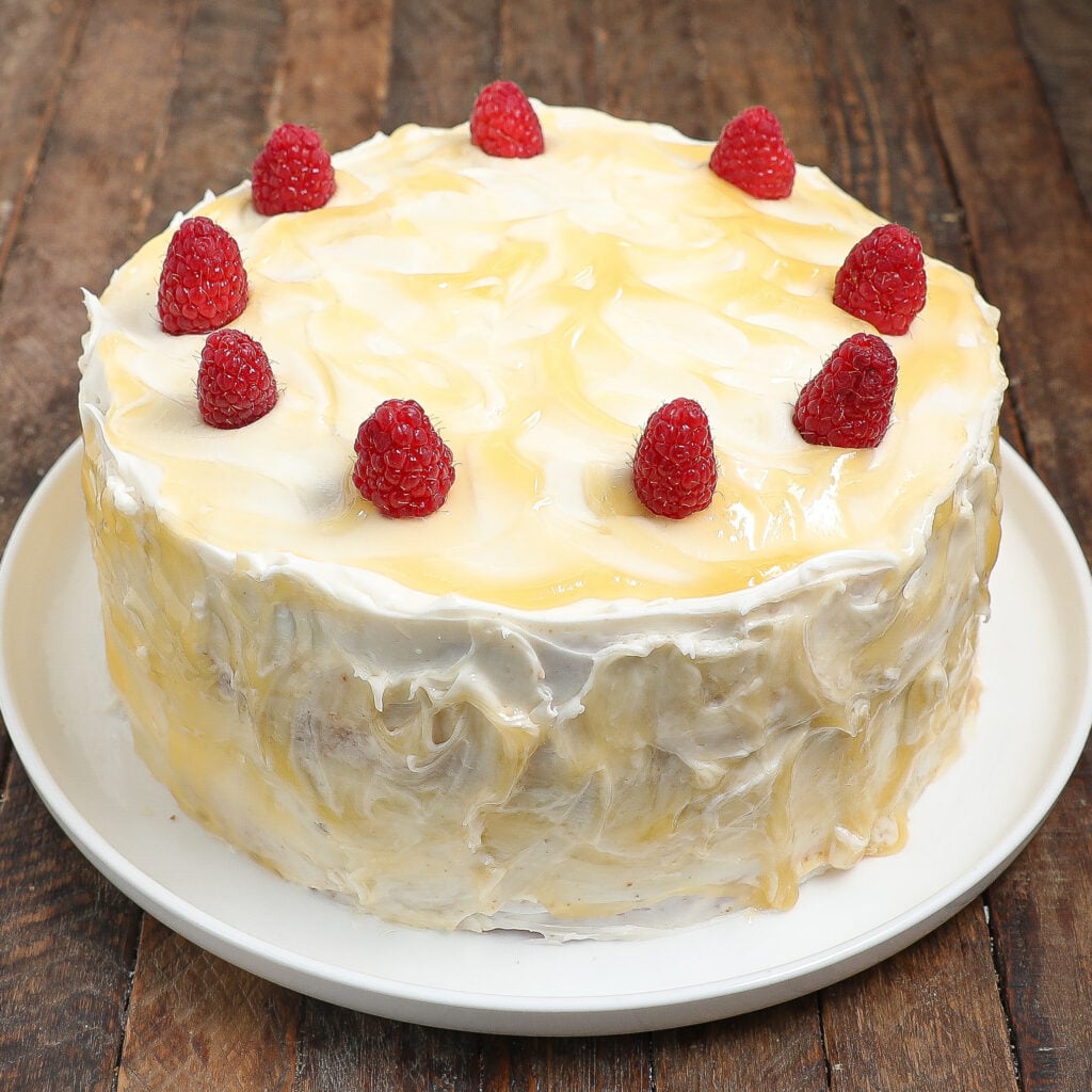 A Raspberry Lemon Cake with white frosting and a light yellow glaze, topped with seven raspberries, sits on a white plate on a wooden surface.