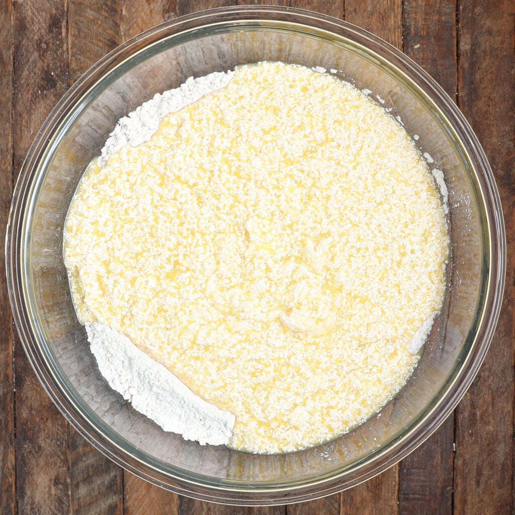 A glass bowl on a wooden surface contains flour and melted butter, partially mixed together, as the first step in preparing a Raspberry Lemon Cake, with the flour still visible around the edges.