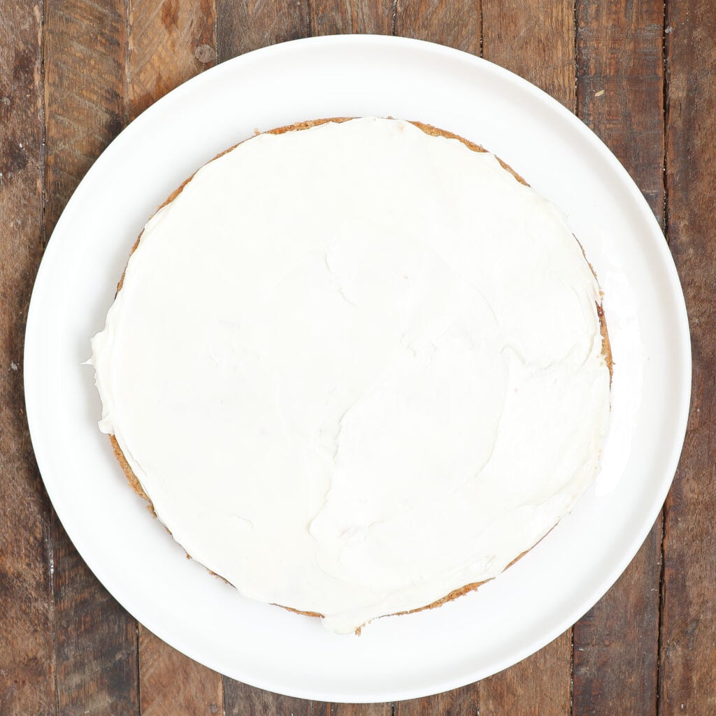 A round Raspberry Lemon Cake covered with smooth white frosting sits on a white plate, placed on a rustic wooden surface.