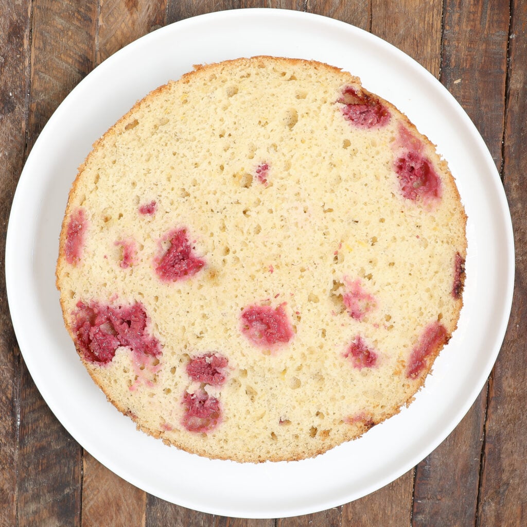 A round, plain Raspberry Lemon Cake with visible raspberry pieces rests on a white plate atop a rustic wooden surface.