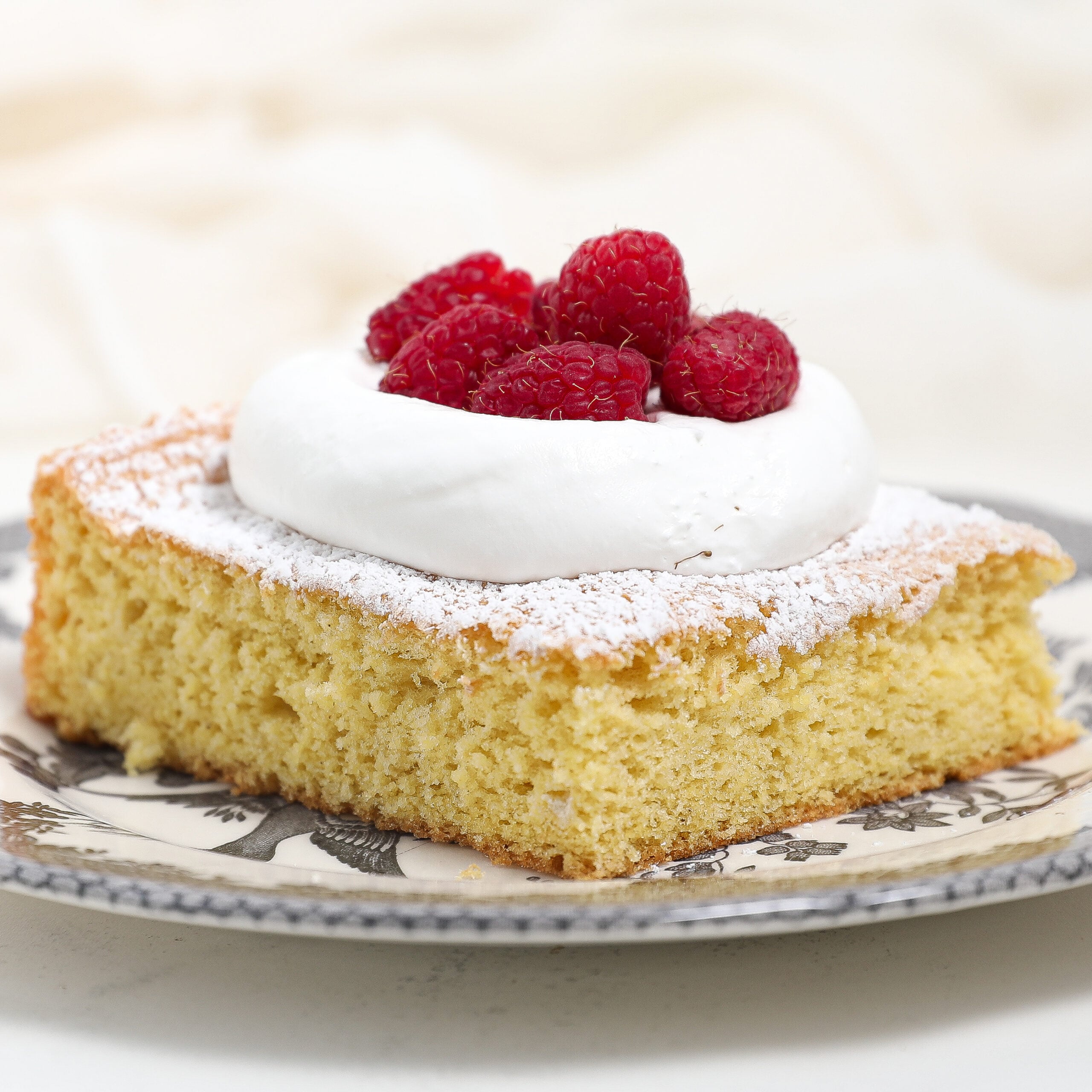 A slice of classic sponge cake topped with a dollop of whipped cream and fresh raspberries, dusted with powdered sugar and served on a decorative plate.