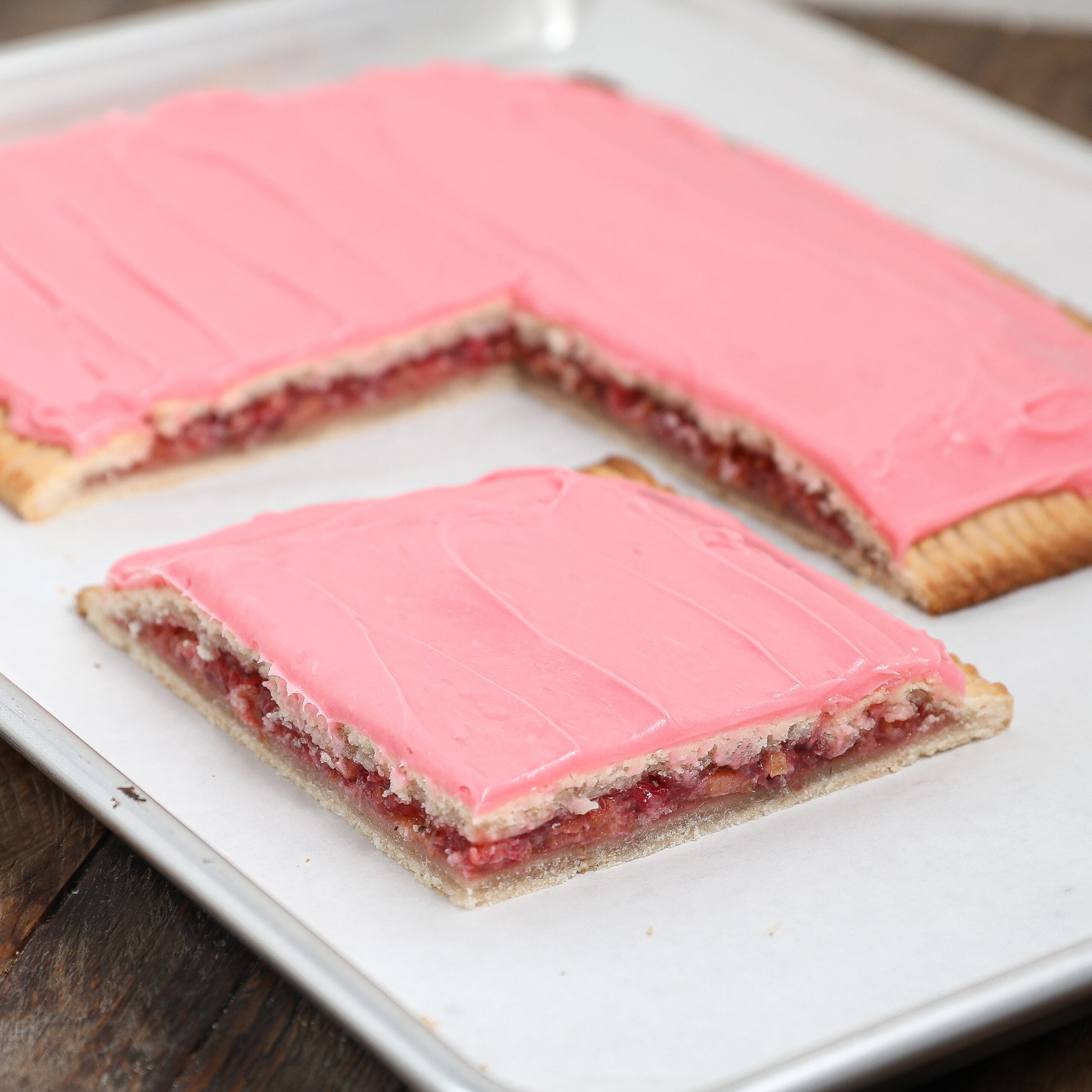 A rectangular Strawberry Rhubarb Pop Tart with pink frosting is cut, revealing a layer of fruit filling inside. One large piece is separated from the rest, all on a parchment-lined baking sheet.