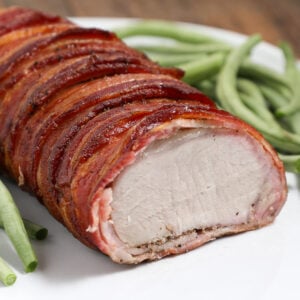 A Bacon-Wrapped Pork Loin is sliced to reveal its juicy, cooked interior, served on a white plate with fresh green beans in the background.