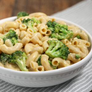 A white bowl filled with Easy Cheesy Broccoli Pasta—creamy macaroni and cheese mixed with bright green broccoli florets—sits atop a striped cloth.