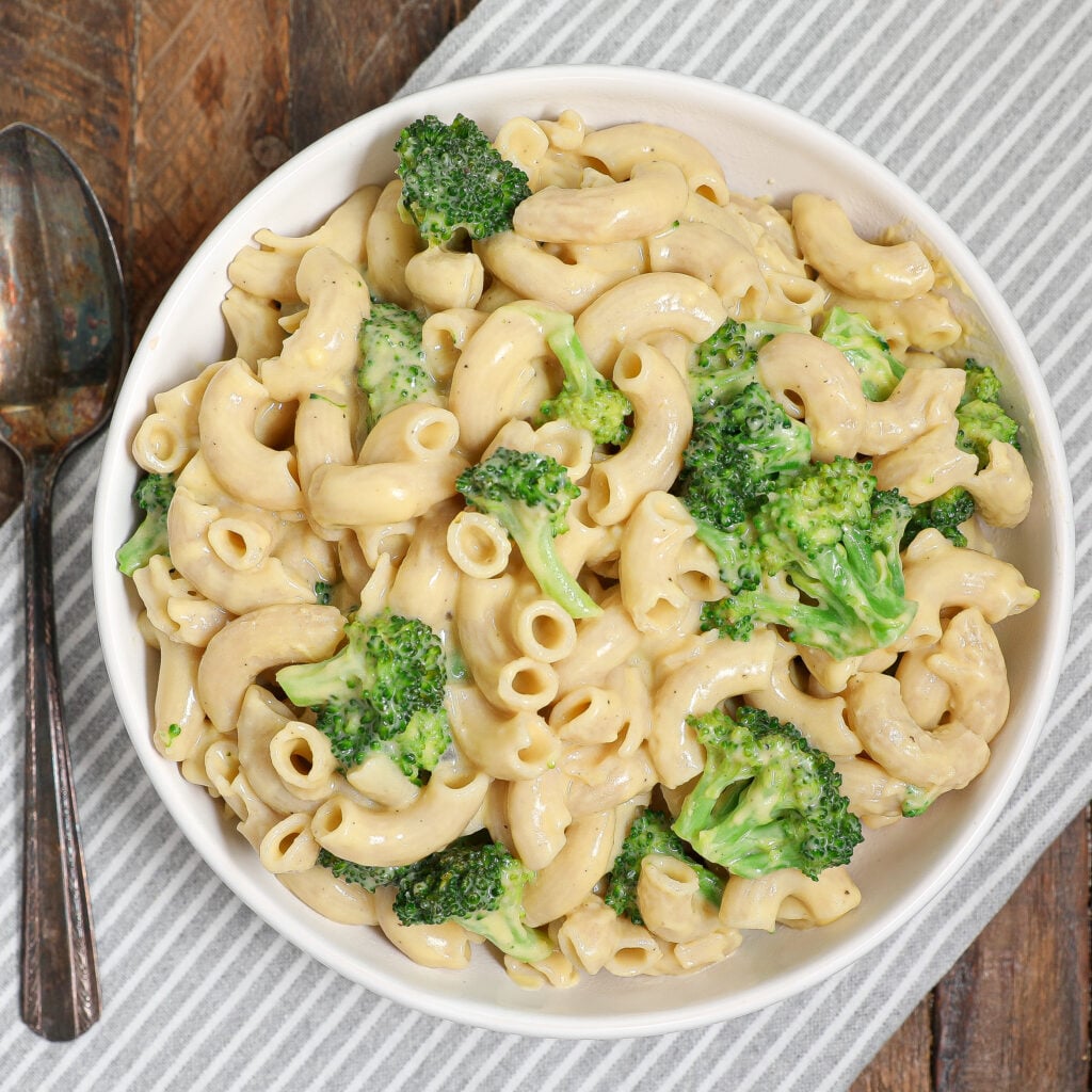 A bowl of Easy Cheesy Broccoli Pasta, with macaroni and broccoli florets, sits on a striped cloth next to two metal spoons on a wooden table.