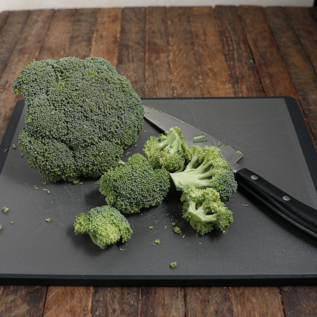 A head of broccoli and several florets sit on a cutting board next to a kitchen knife, ready to be chopped for Easy Cheesy Broccoli Pasta, all placed on a wooden table.