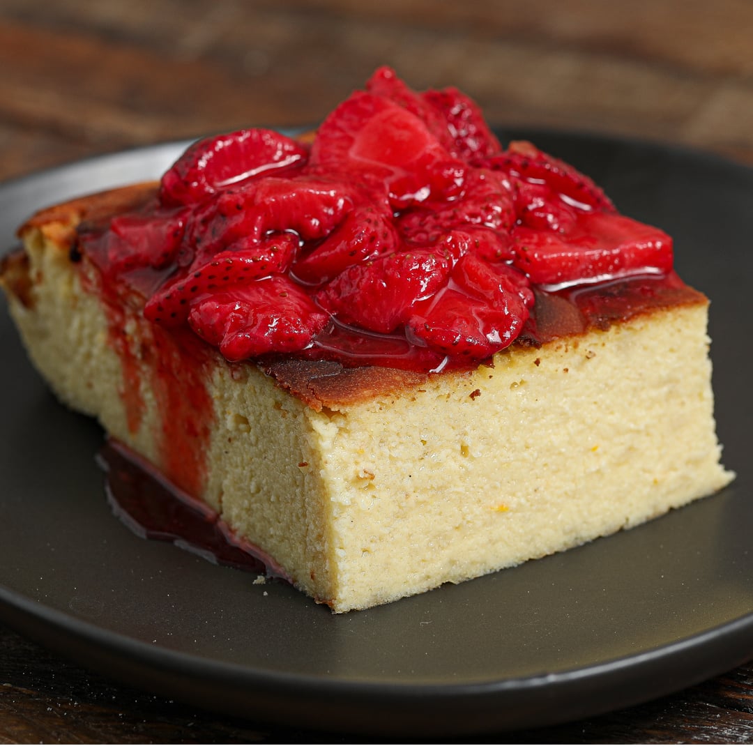 A slice of Basque Cheesecake with Raspberry Sauce sits on a dark plate, topped with a generous amount of glossy, syrupy strawberries and placed on a wooden surface.