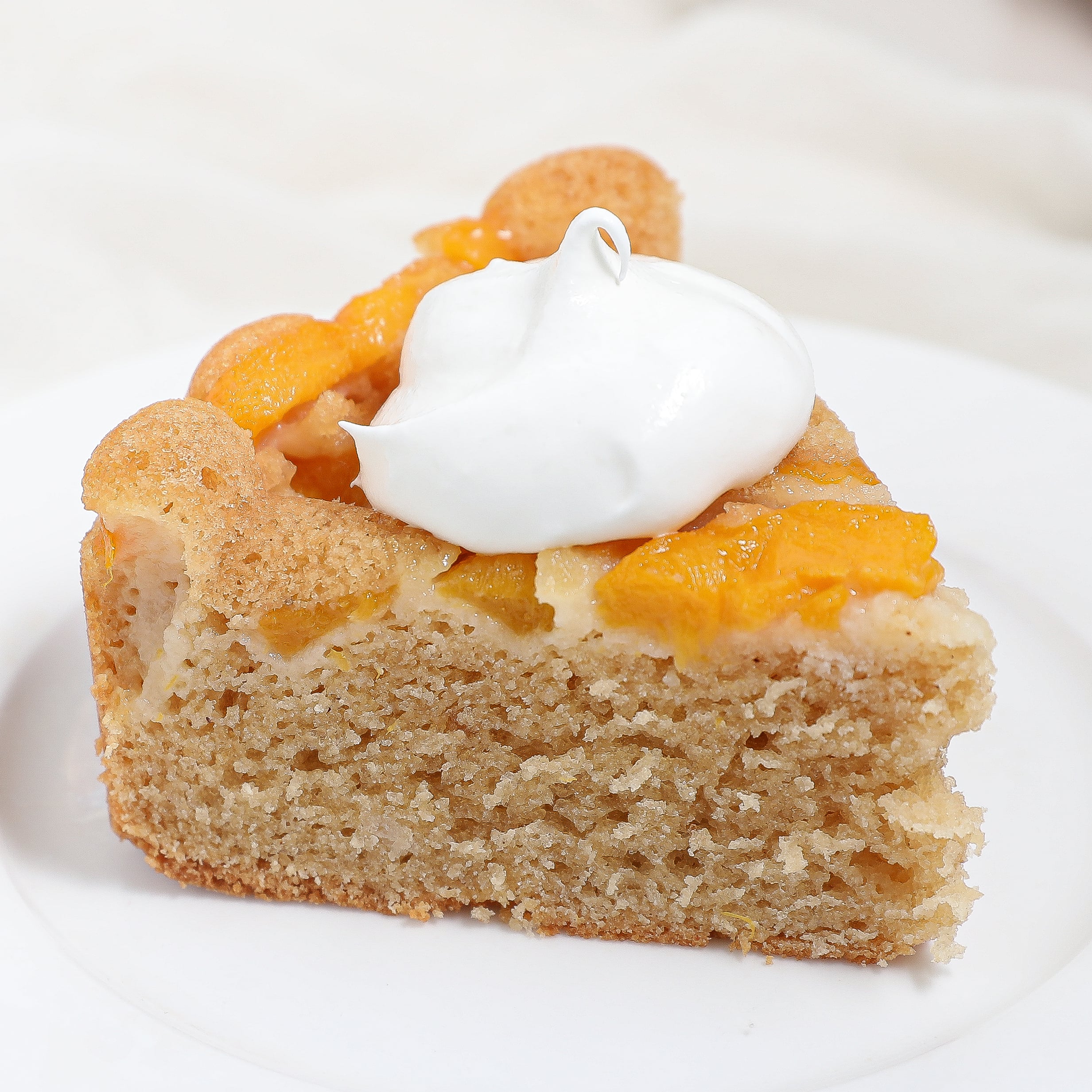 A slice of peach cake topped with a dollop of whipped cream, served on a white plate for a delightful dessert.
